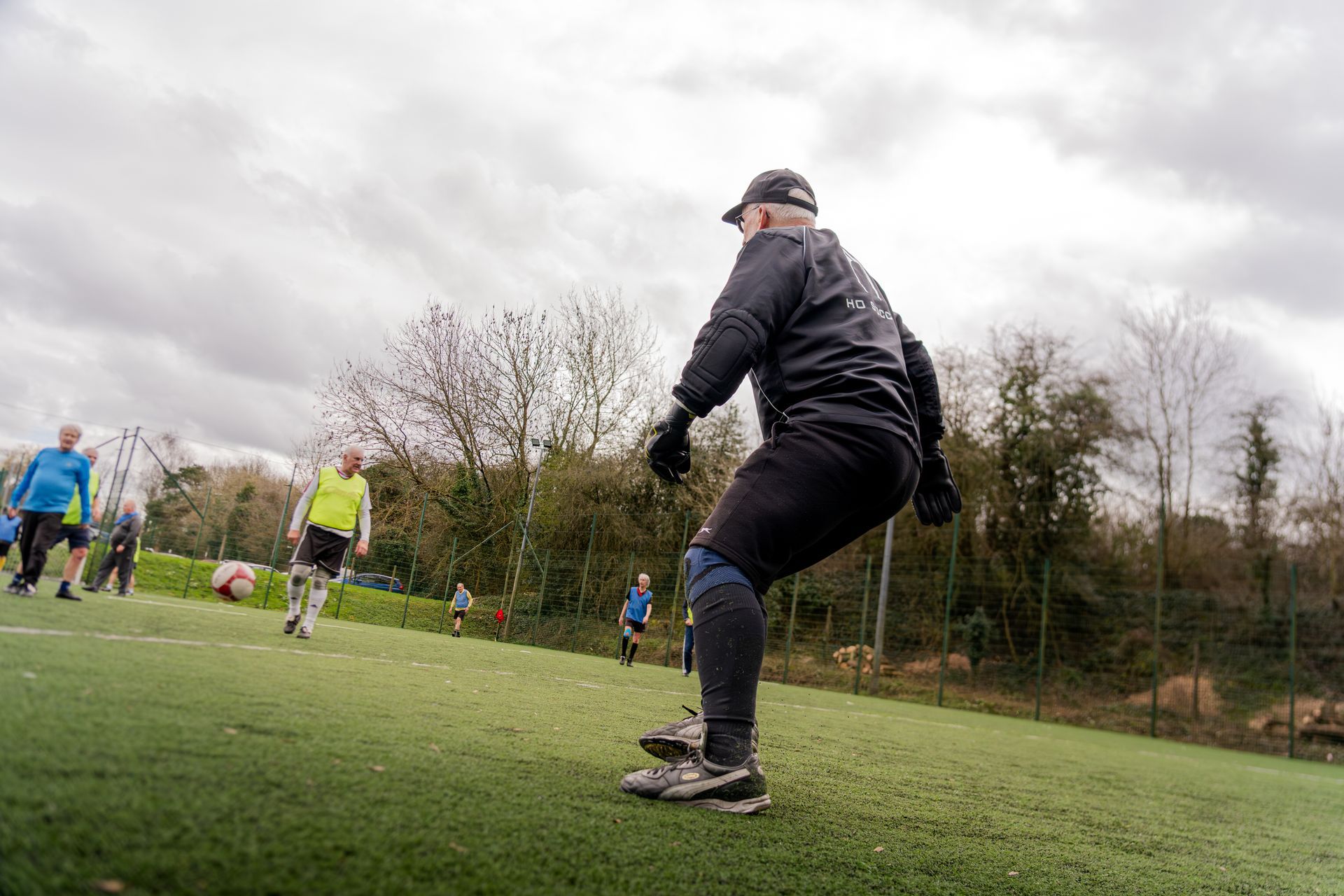 Person in black sportswear crouches on a soccer field as players practice in the background.