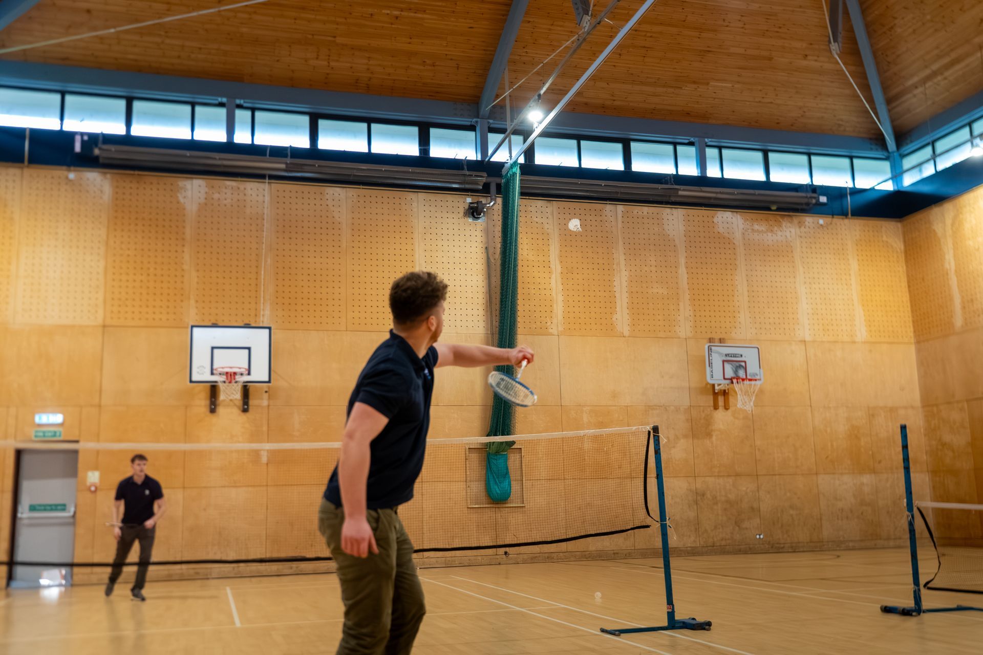 Indoor gym with two people playing badminton near a net and wooden walls