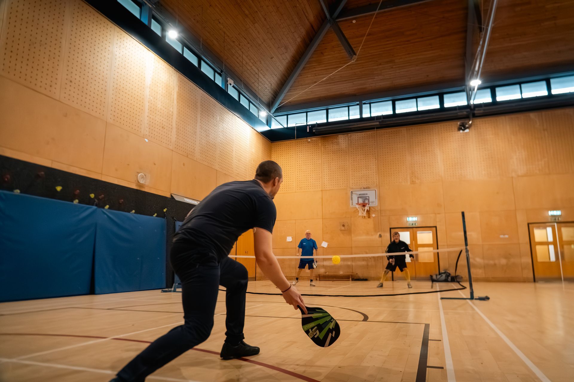 Indoor pickleball game in a gym, with players and a man in black serving in the foreground