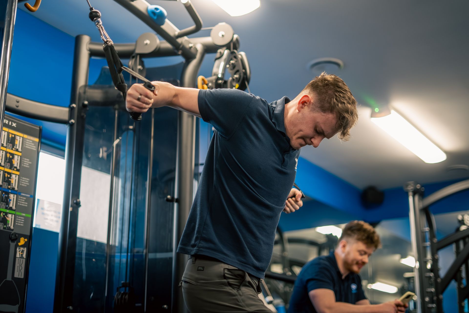 Man using a cable machine in a blue gym, with another person exercising in the background.