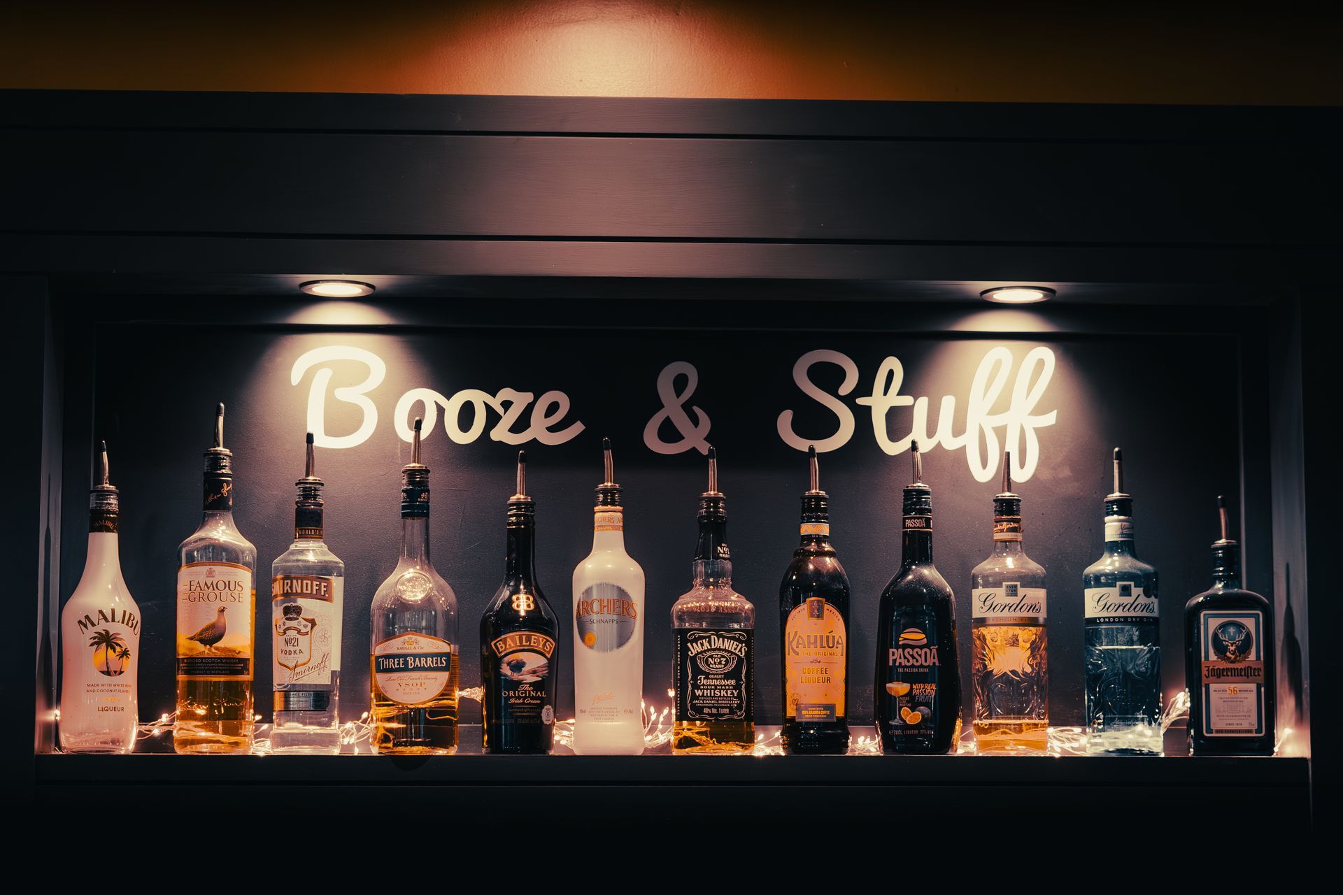 Bar shelf with assorted liquor bottles under neon “Booze & Stuff” sign