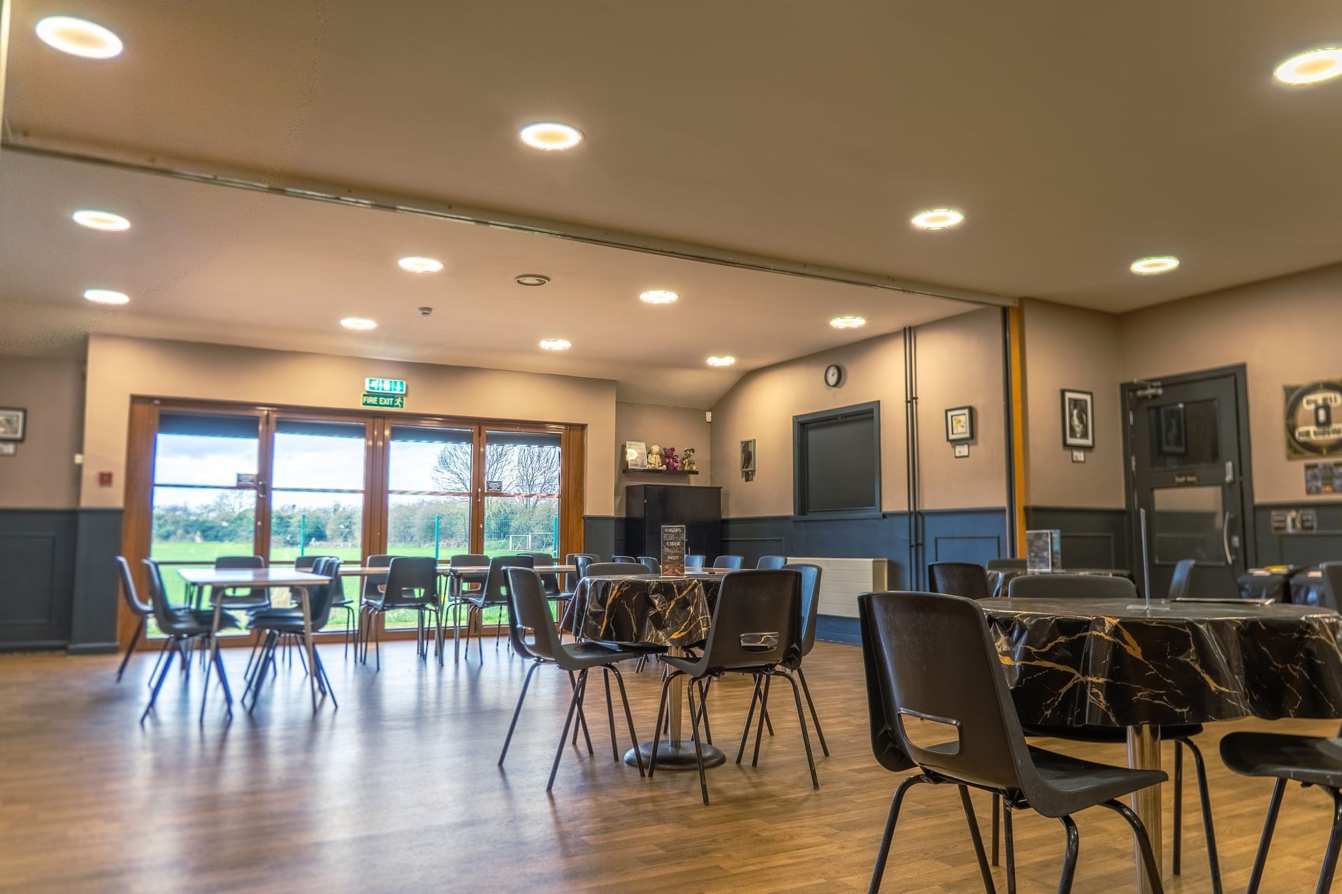 Empty dining hall with tables and chairs, wooden floor, recessed lights, and glass doors opening outside