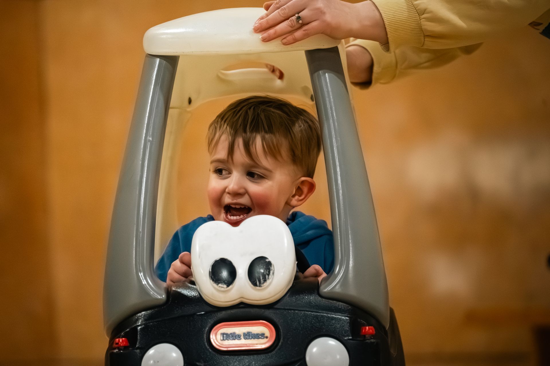 Child smiling in a toy car at a playground, with an adult’s hand steadying the canopy.