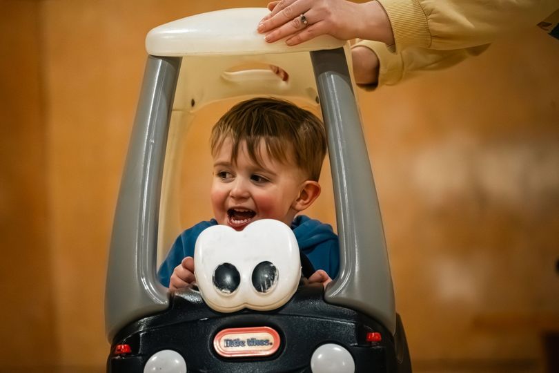 Toddler smiling in a toy car while an adult hand steadies the top handle indoors