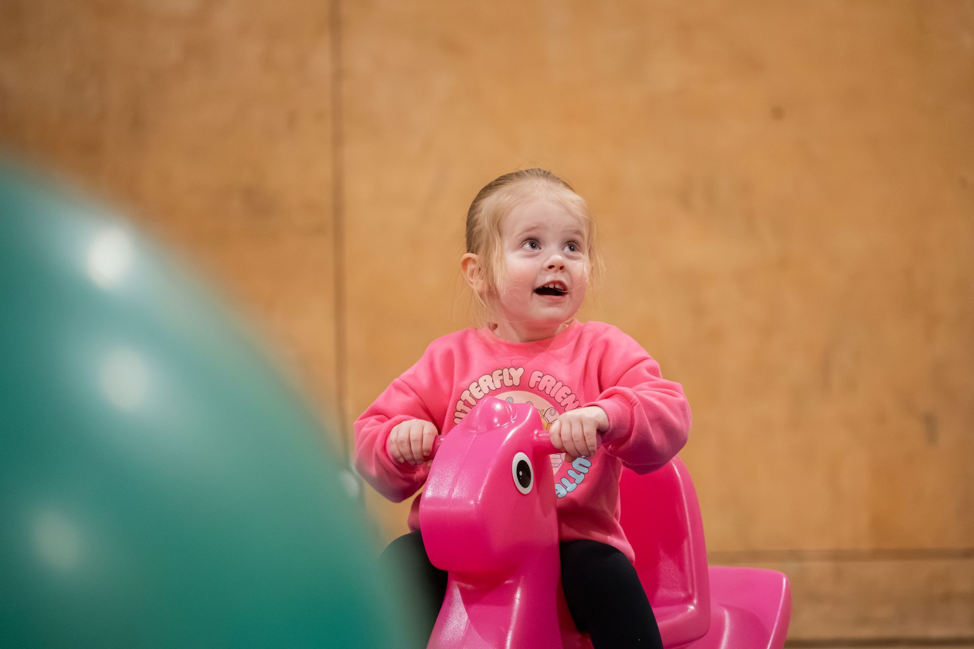 Child on a pink toy ride, smiling and holding the handlebars indoors