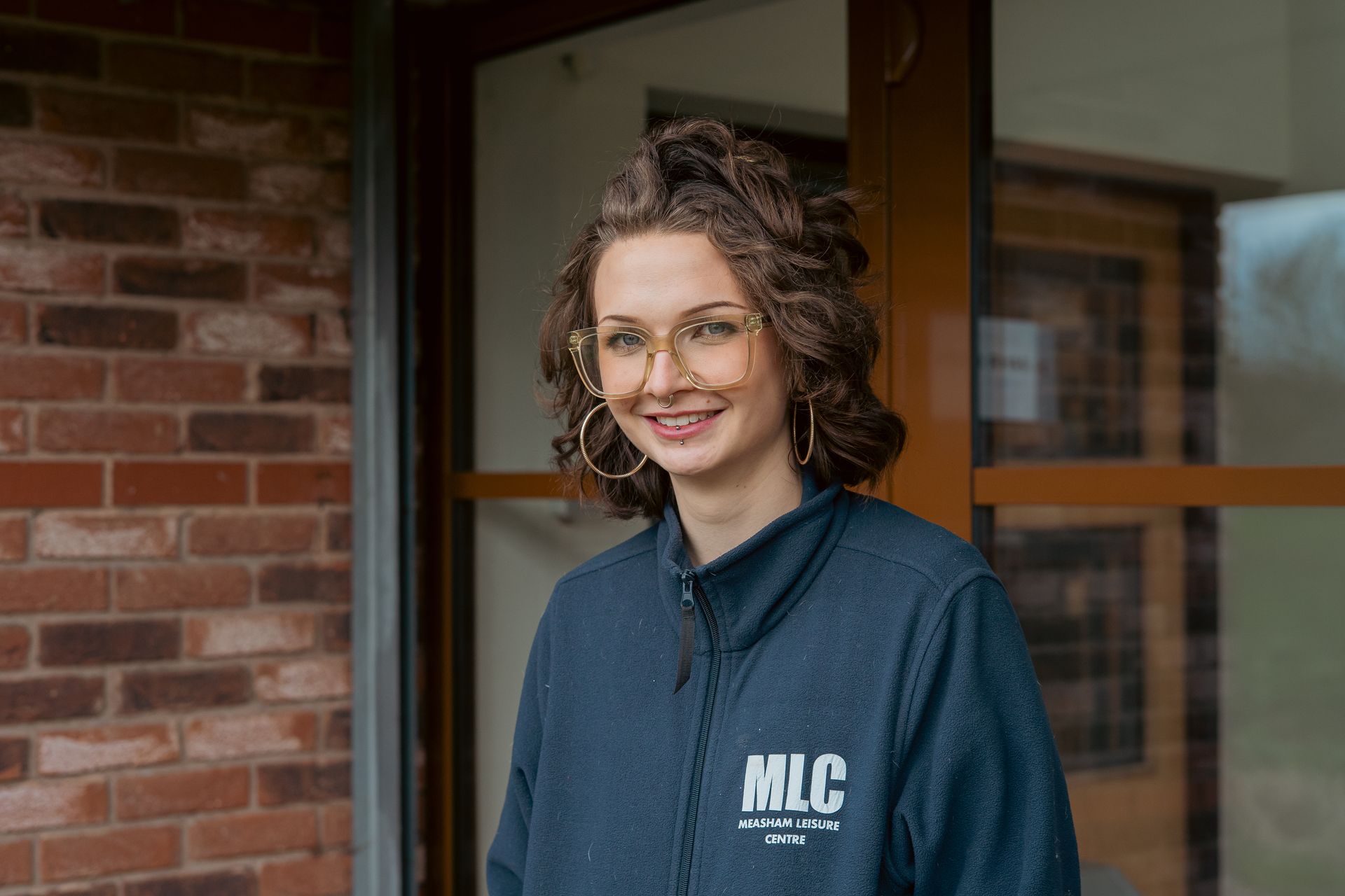 Smiling person in glasses and a dark MLC jacket standing by a brick wall and glass door