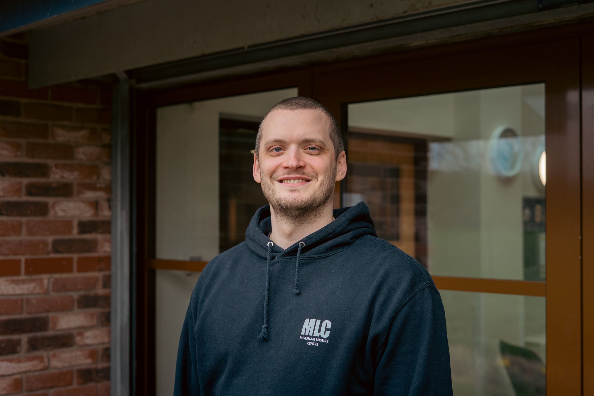 Man smiling in a dark hoodie standing outside a brick building with glass doors