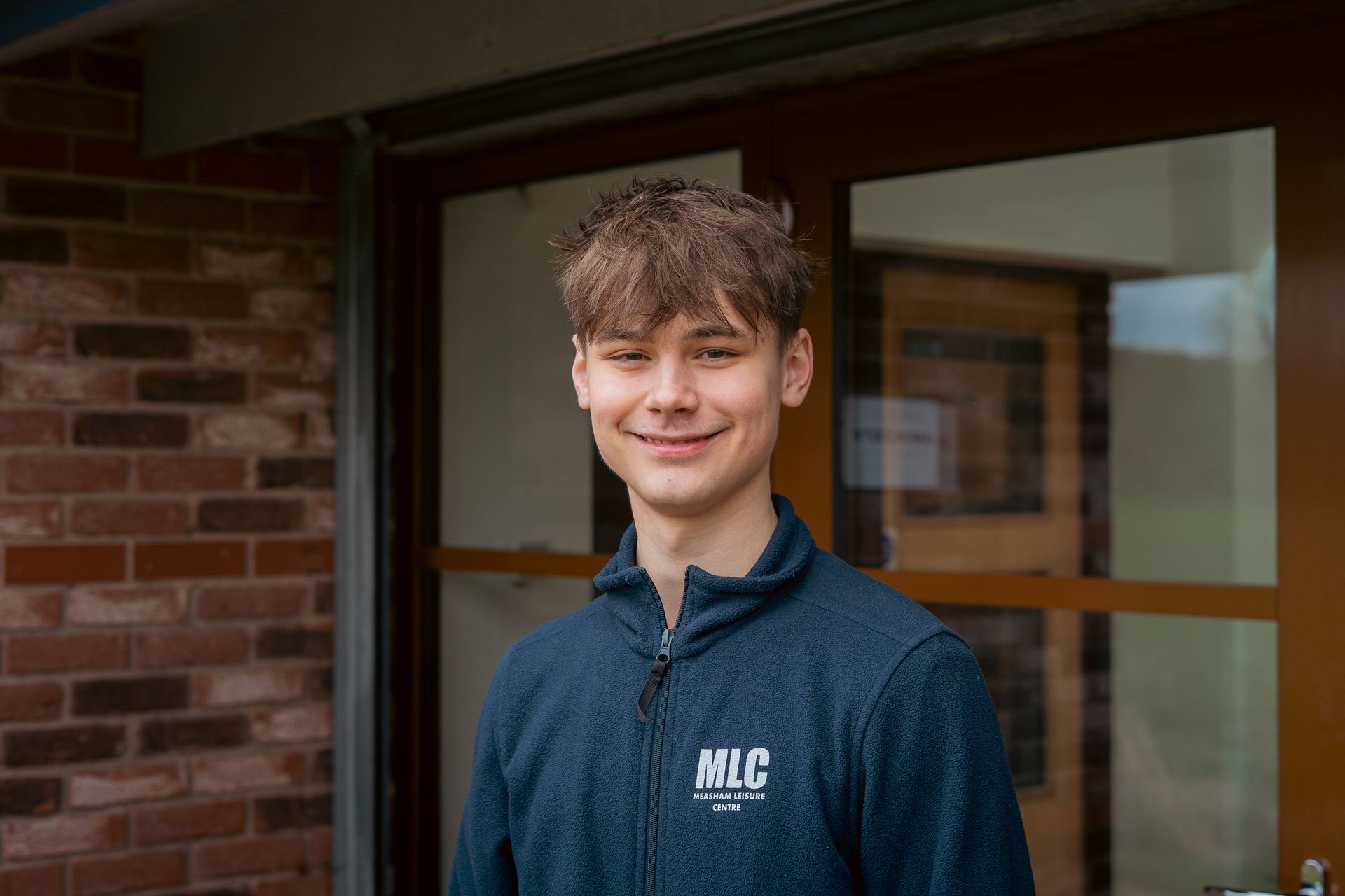 Young person in a blue sweater smiling in front of a brick building entrance