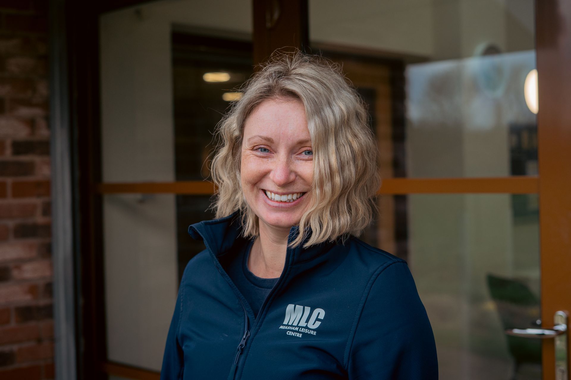 Smiling person in a navy jacket standing indoors by a glass door
