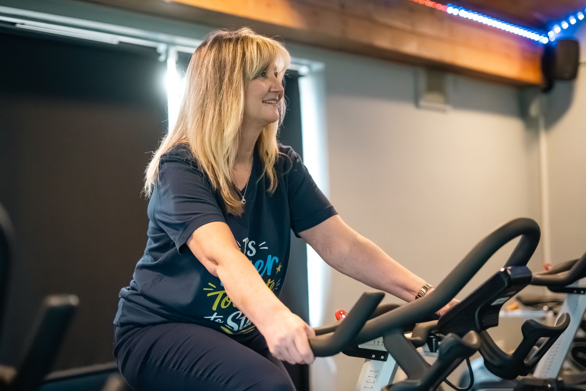 Woman exercising on a stationary bike in a gym, wearing a dark T-shirt and smiling.
