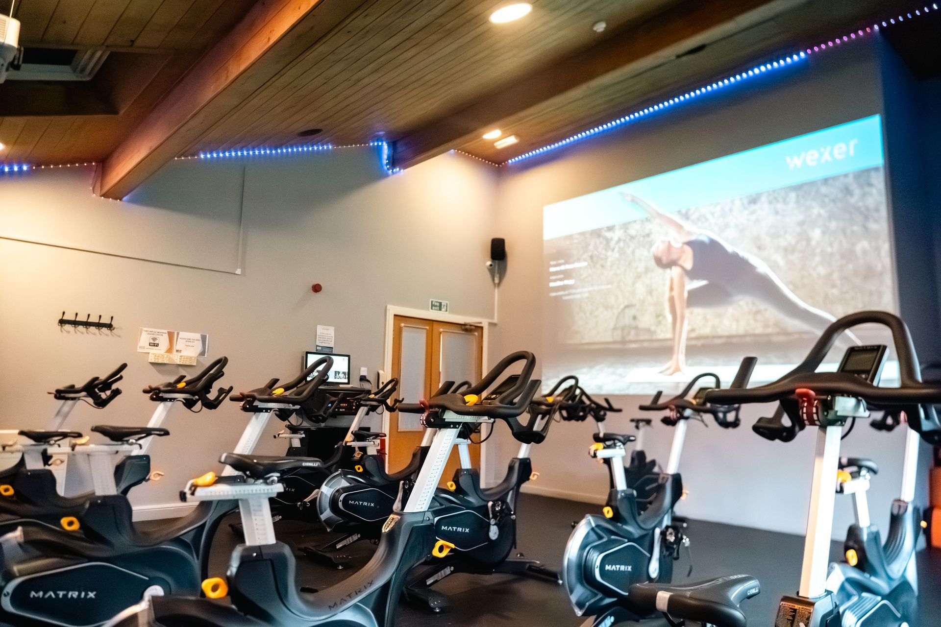 Indoor cycling studio with stationary bikes and a large wall projector showing a workout scene