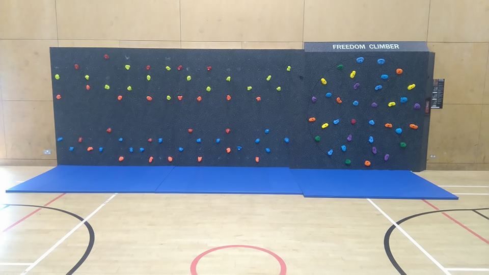 Indoor climbing wall with colorful holds above a blue padded mat in a gymnasium.