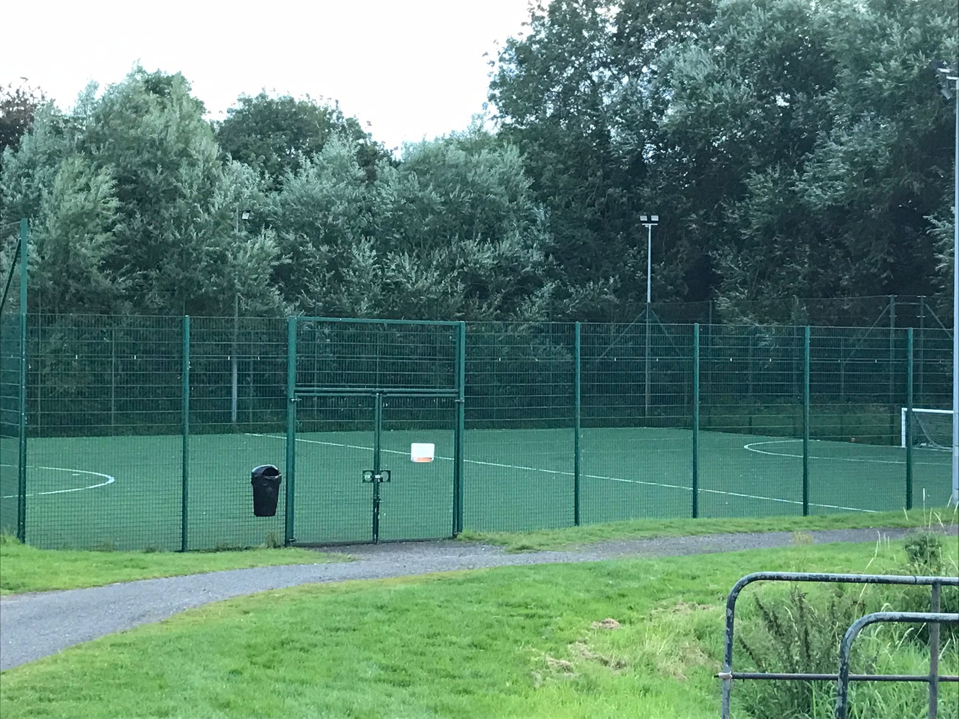 Empty fenced sports field beside a grassy path, with trees and cloudy sky in the background