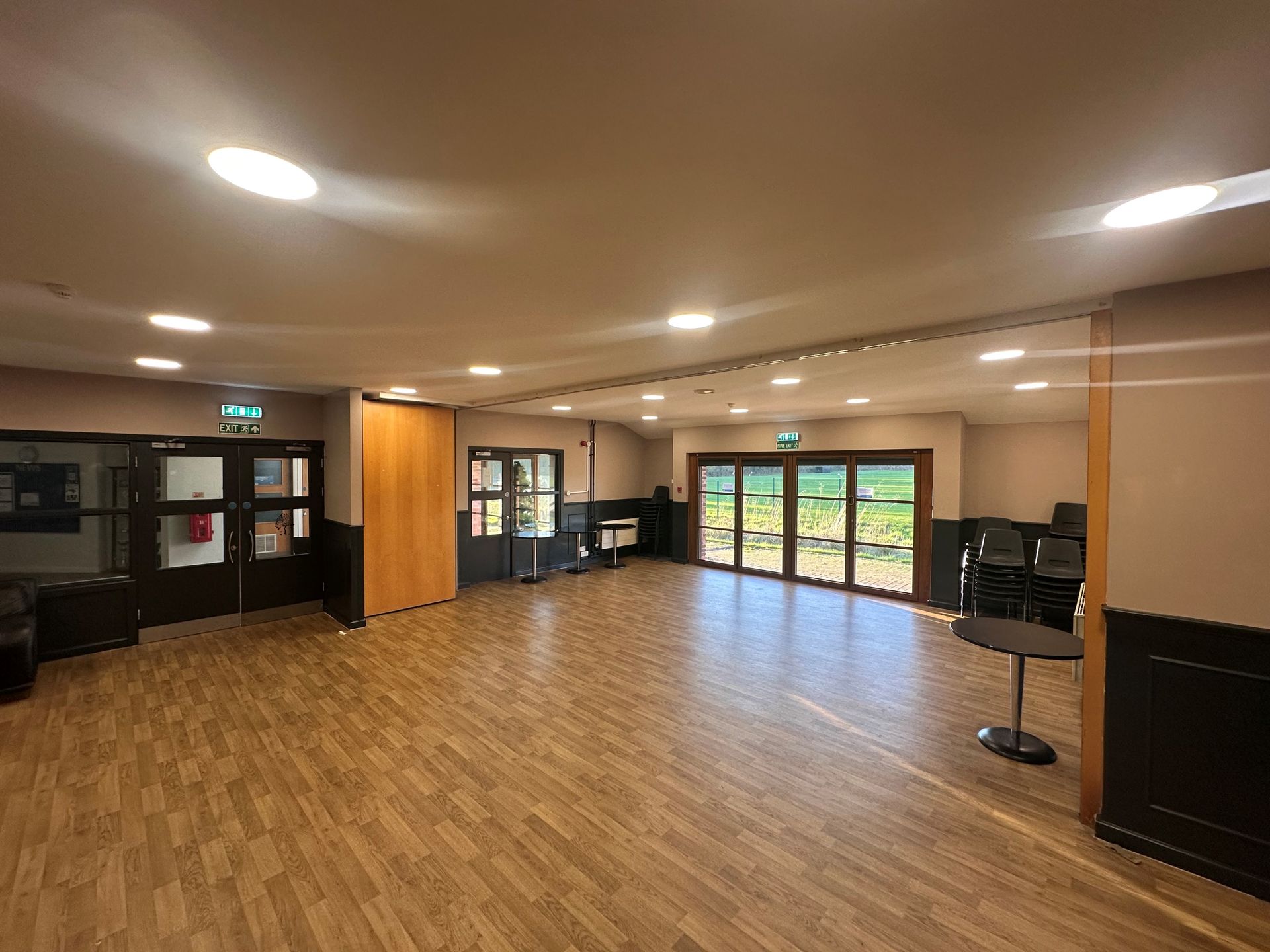 Empty community hall with wood floor, ceiling lights, and glass doors opening to greenery.
