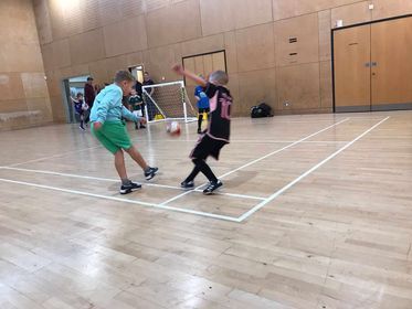Two children play badminton in a gymnasium, with a net and wooden walls in the background.