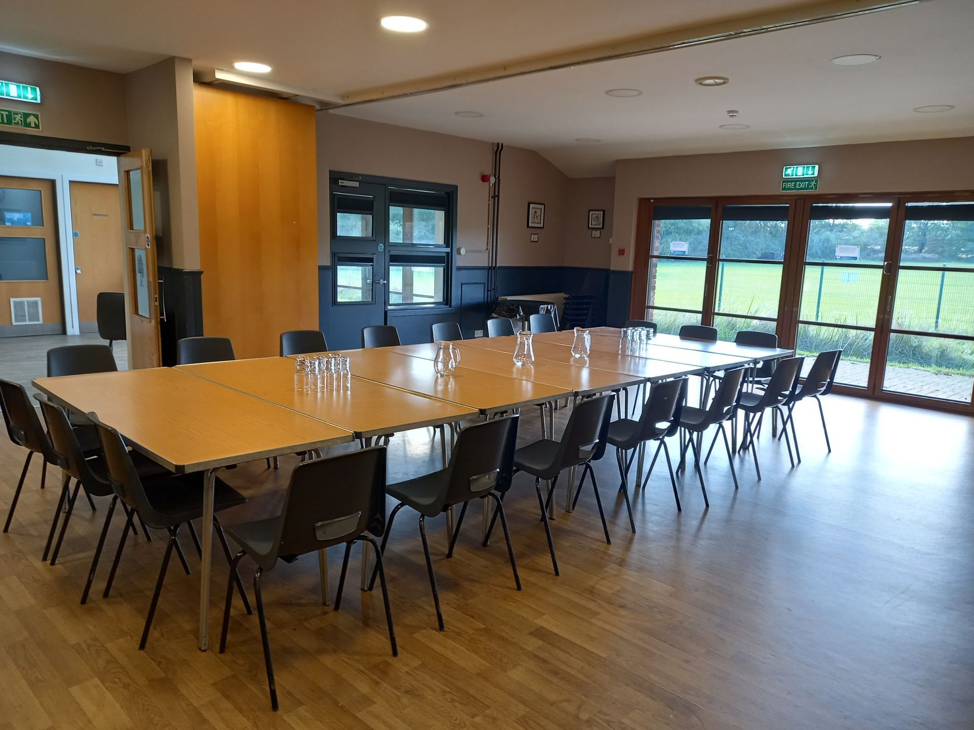 Empty conference room with long wooden table, black chairs, and large windows overlooking greenery.
