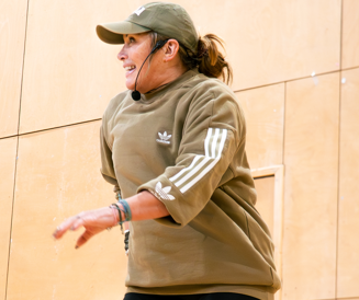 Person in a tan hoodie and cap gesturing in a gym-like room against a wooden wall