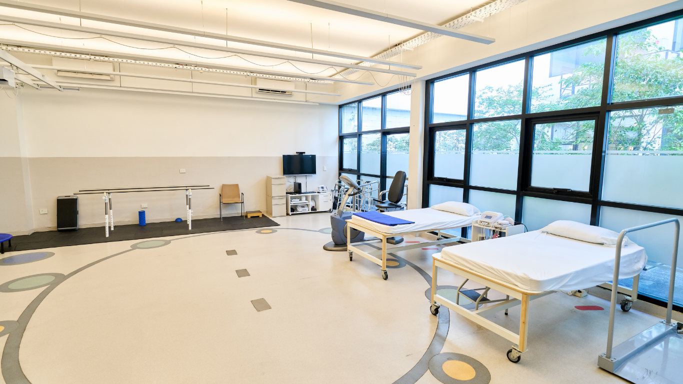 Bright rehabilitation room with treatment tables, equipment, and large windows