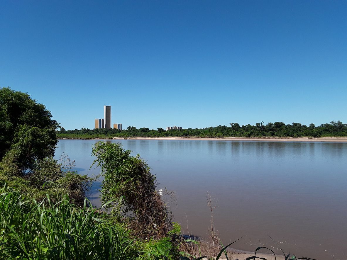 Calm river with blue sky, green shoreline, and distant city towers on the horizon