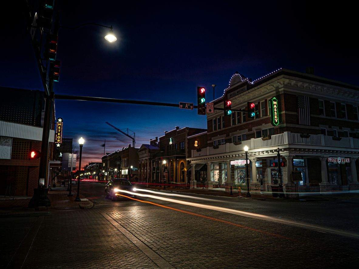 Nighttime city street with lit storefronts, traffic lights, and light trails under a bright moon