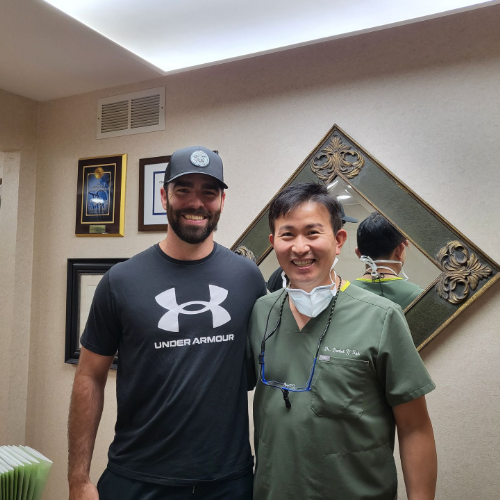 Two smiling men pose indoors, one in a black Under Armour shirt and cap, the other in green scrubs.