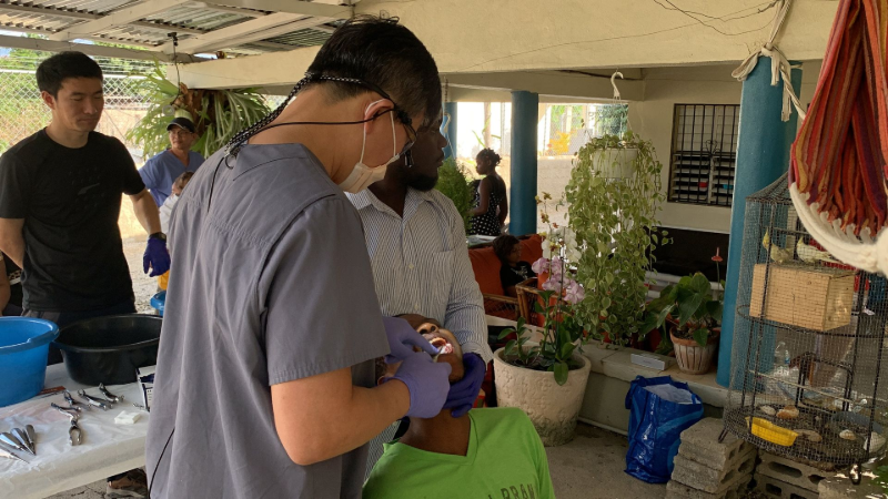 People outdoors under a covered area, one person wearing gloves working at a table with plants and buckets nearby
