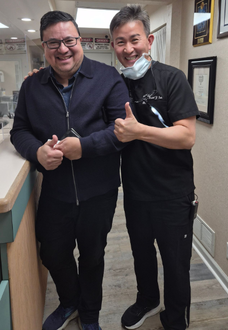 Two smiling people standing in a hallway, giving thumbs up and posing together.