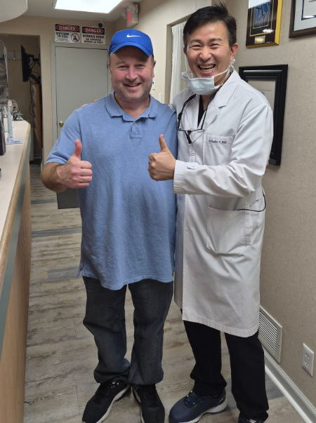 Two smiling men in a hallway, one in a blue cap giving thumbs up, the other in a white lab coat with a mask under chin