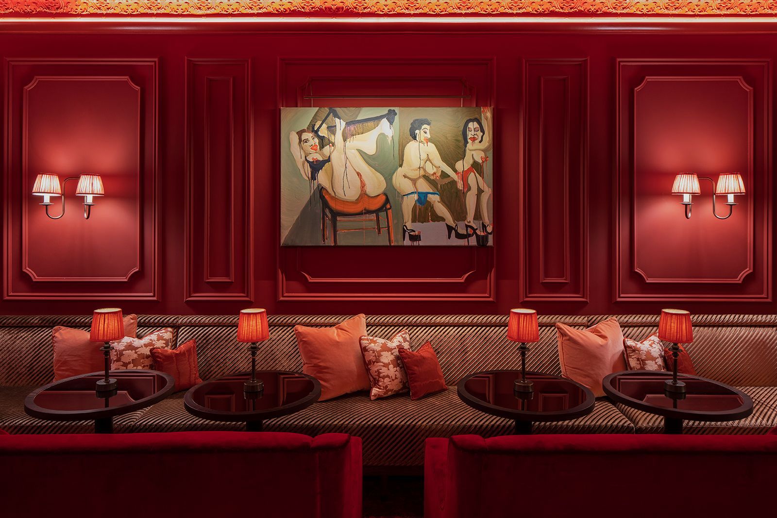 Red booth seating in a bar with a painting, small tables, and lamps.