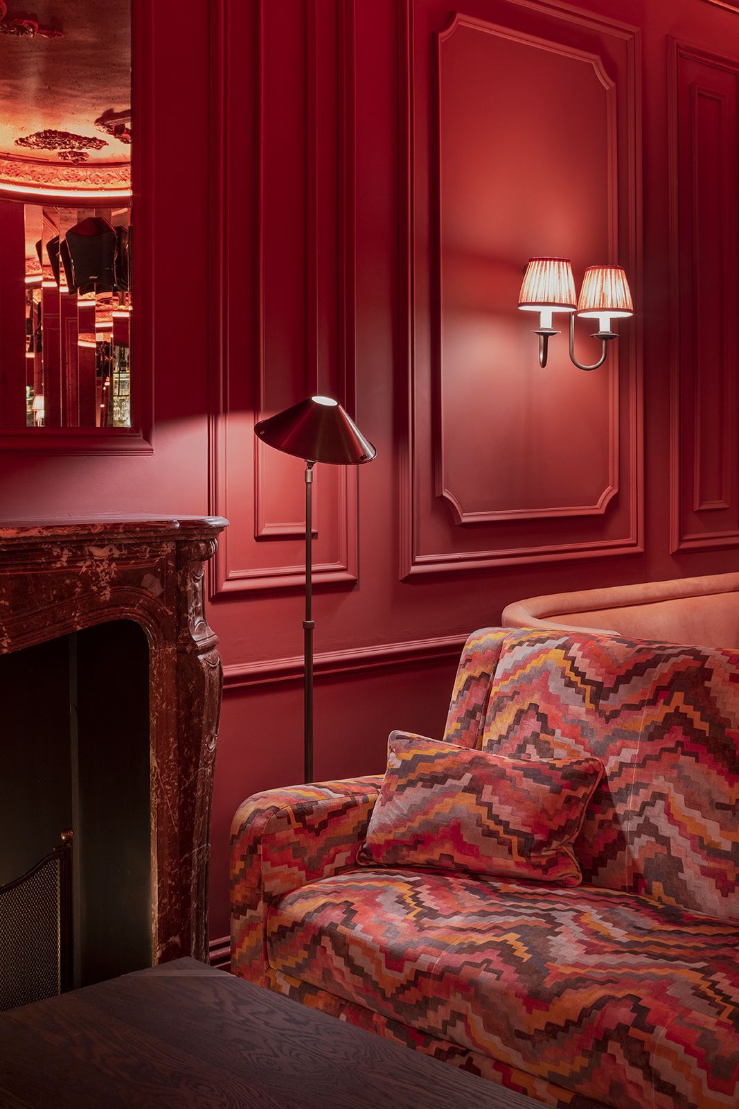 Red-toned room with patterned sofa, floor lamp, and wall sconce. Ornate fireplace and mirror.