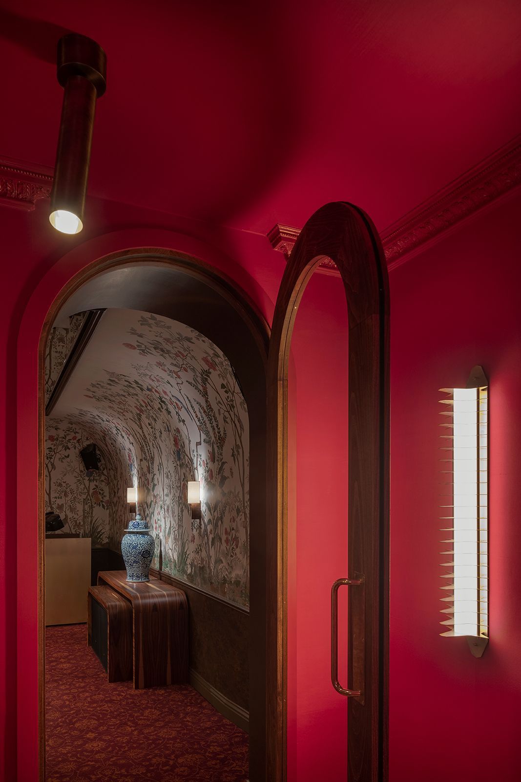 Red hallway with arched doorway; ornate ceiling. Through door, patterned wallpaper and furniture are visible.