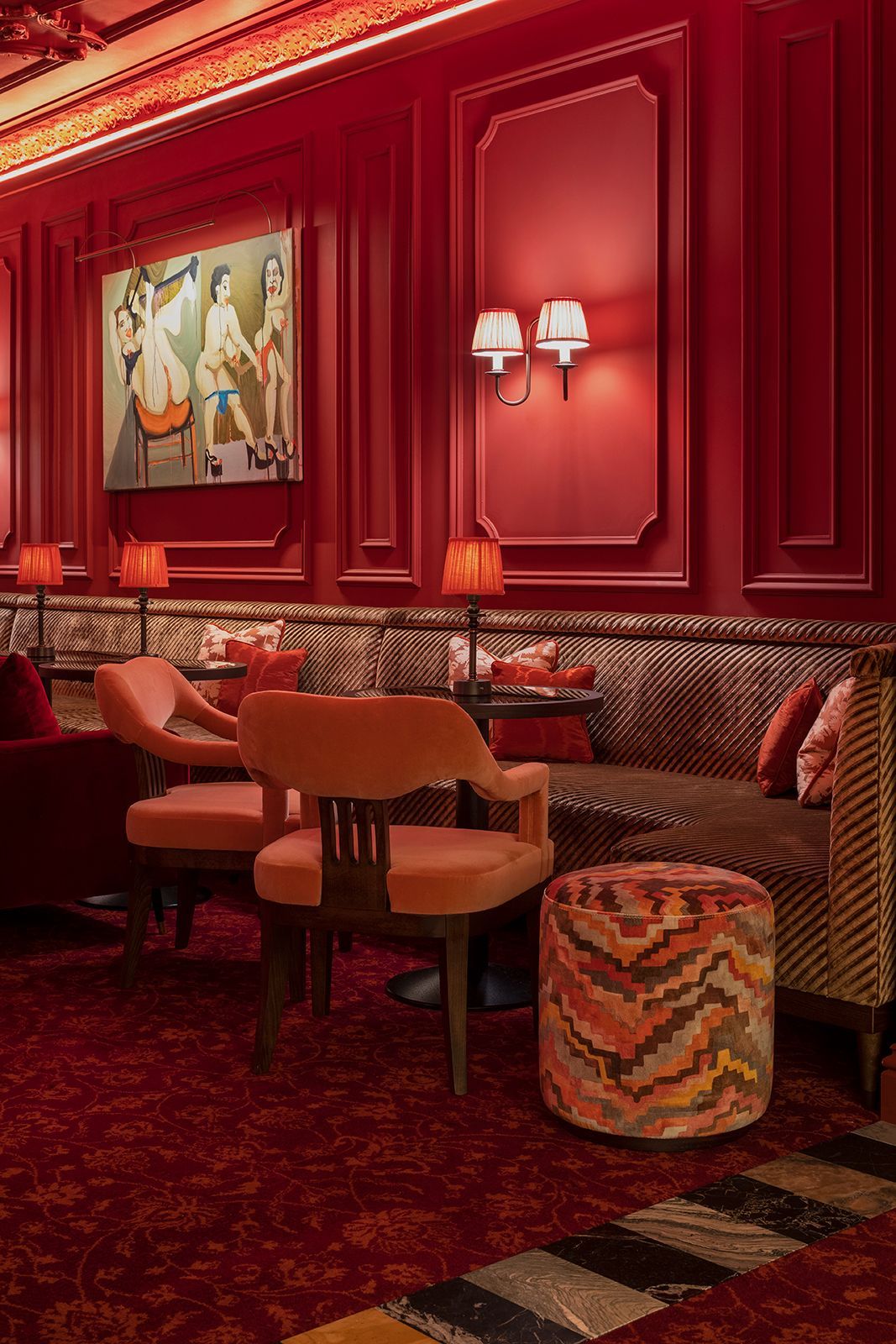 Red-walled lounge with velvet seating, patterned ottoman, lamps, and artwork.