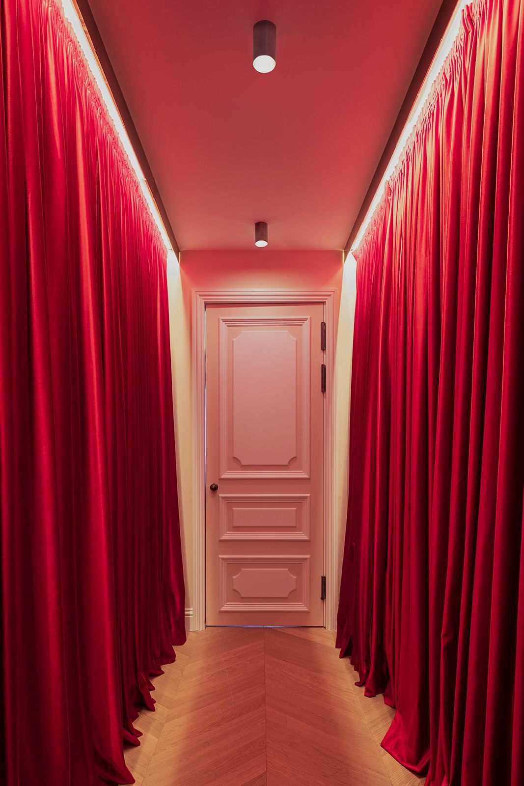 Red velvet curtains line a hallway, leading to a pink door, lit with a warm glow.