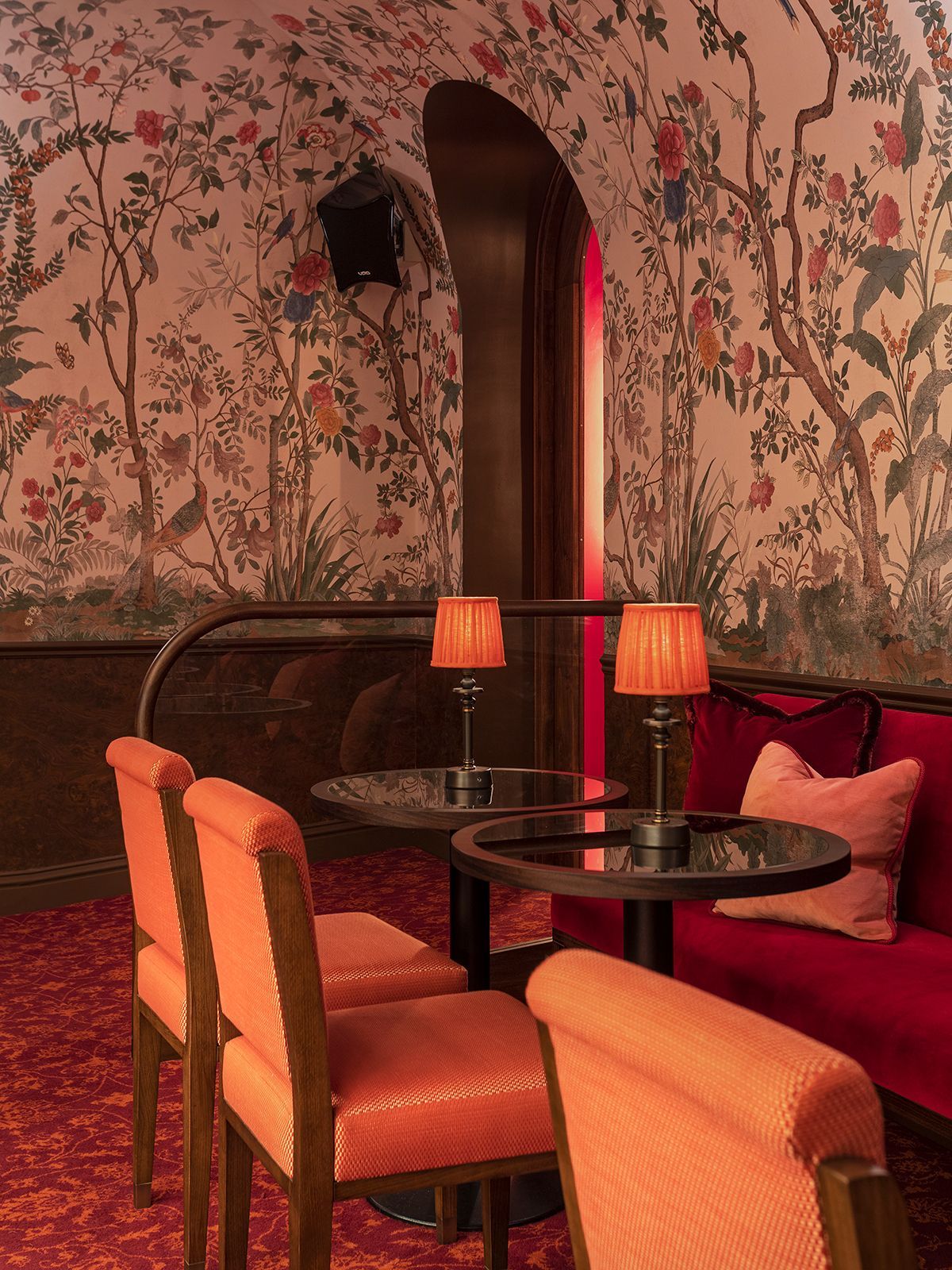 A cozy, reddish-toned bar seating area with floral wallpaper, small tables, orange chairs, and a red sofa.
