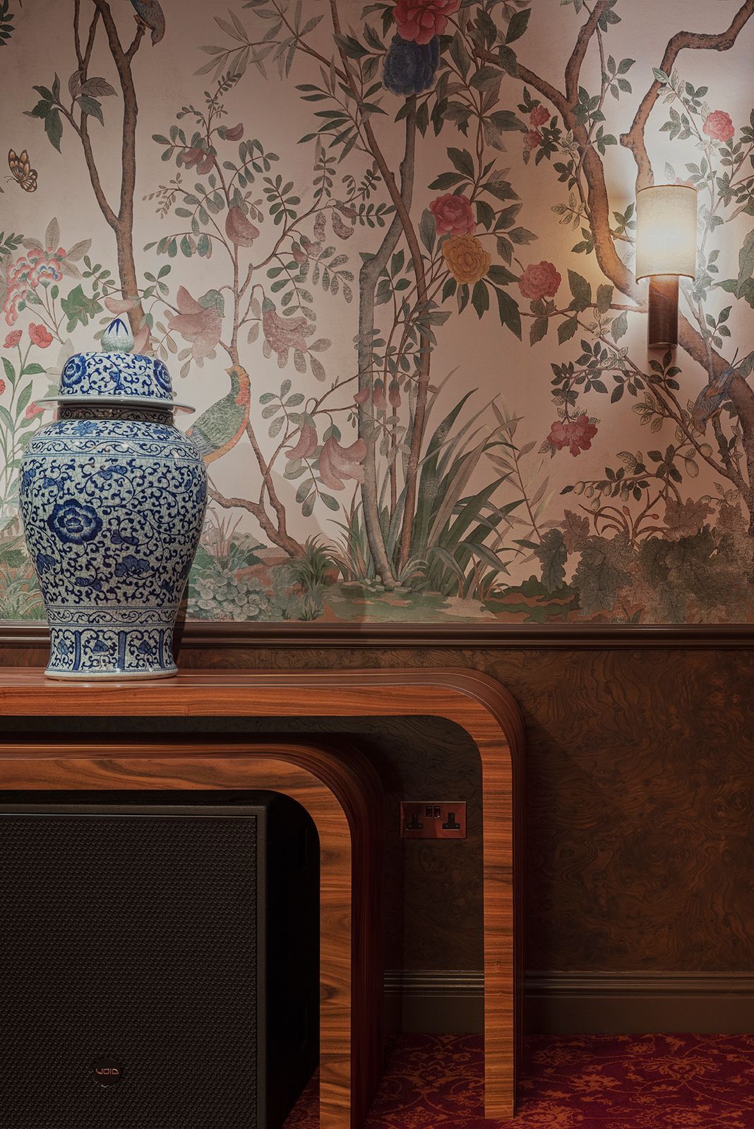 A blue and white porcelain vase sits on a wooden table against floral wallpaper with a wall sconce.