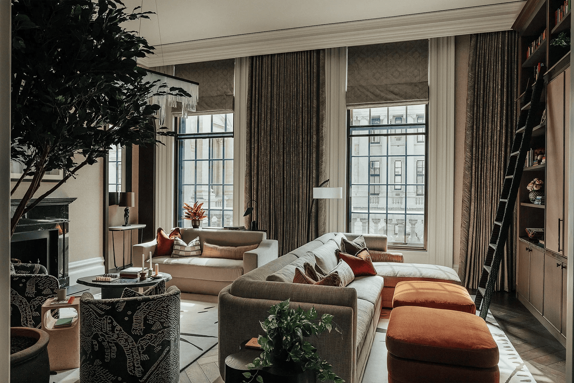 Living room with large windows, tan sofa, orange ottomans, dark drapes, and a tall bookshelf.