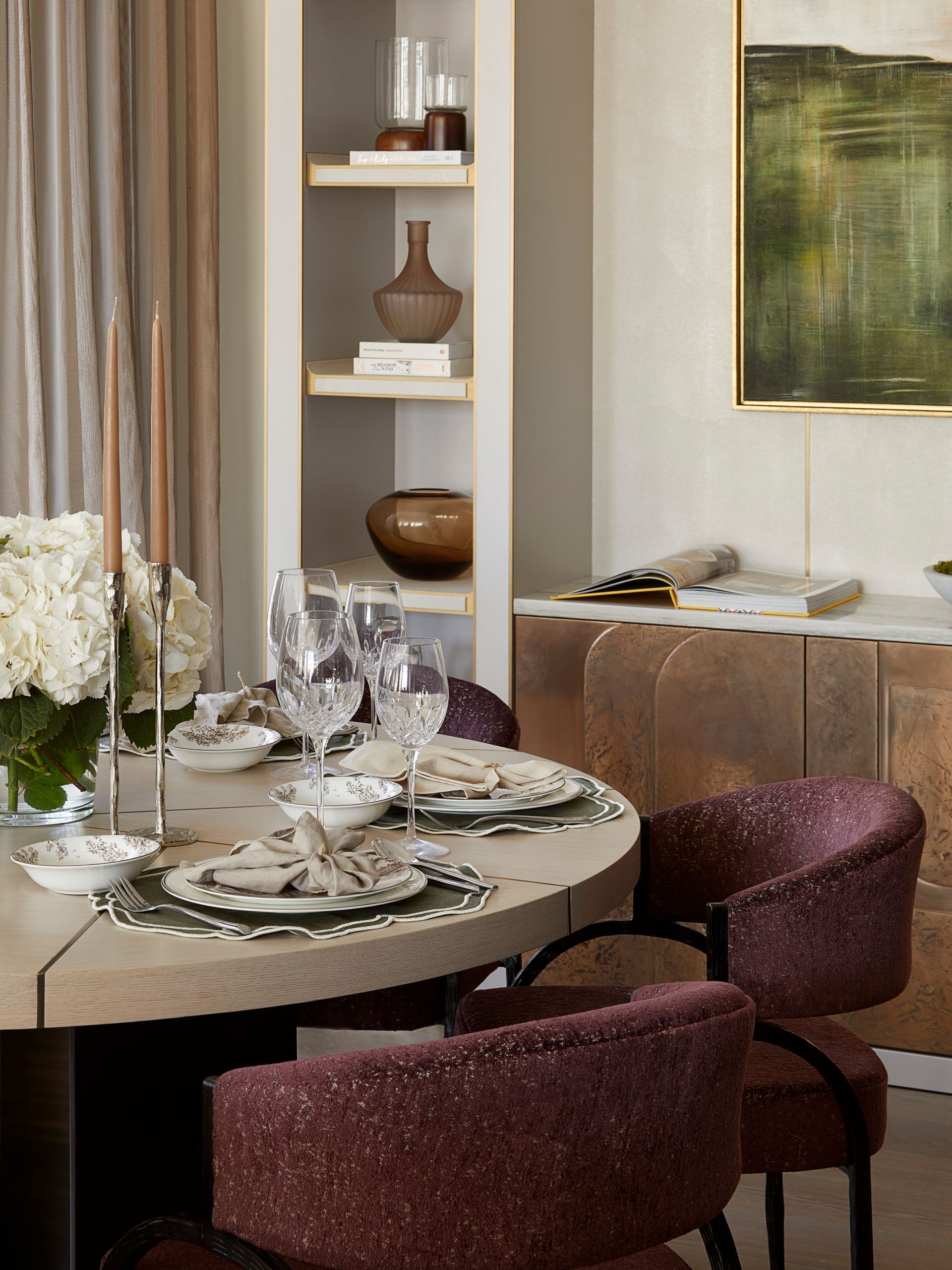 Elegant dining area with round table set for a meal, burgundy chairs, built-in shelves, and a large abstract painting.