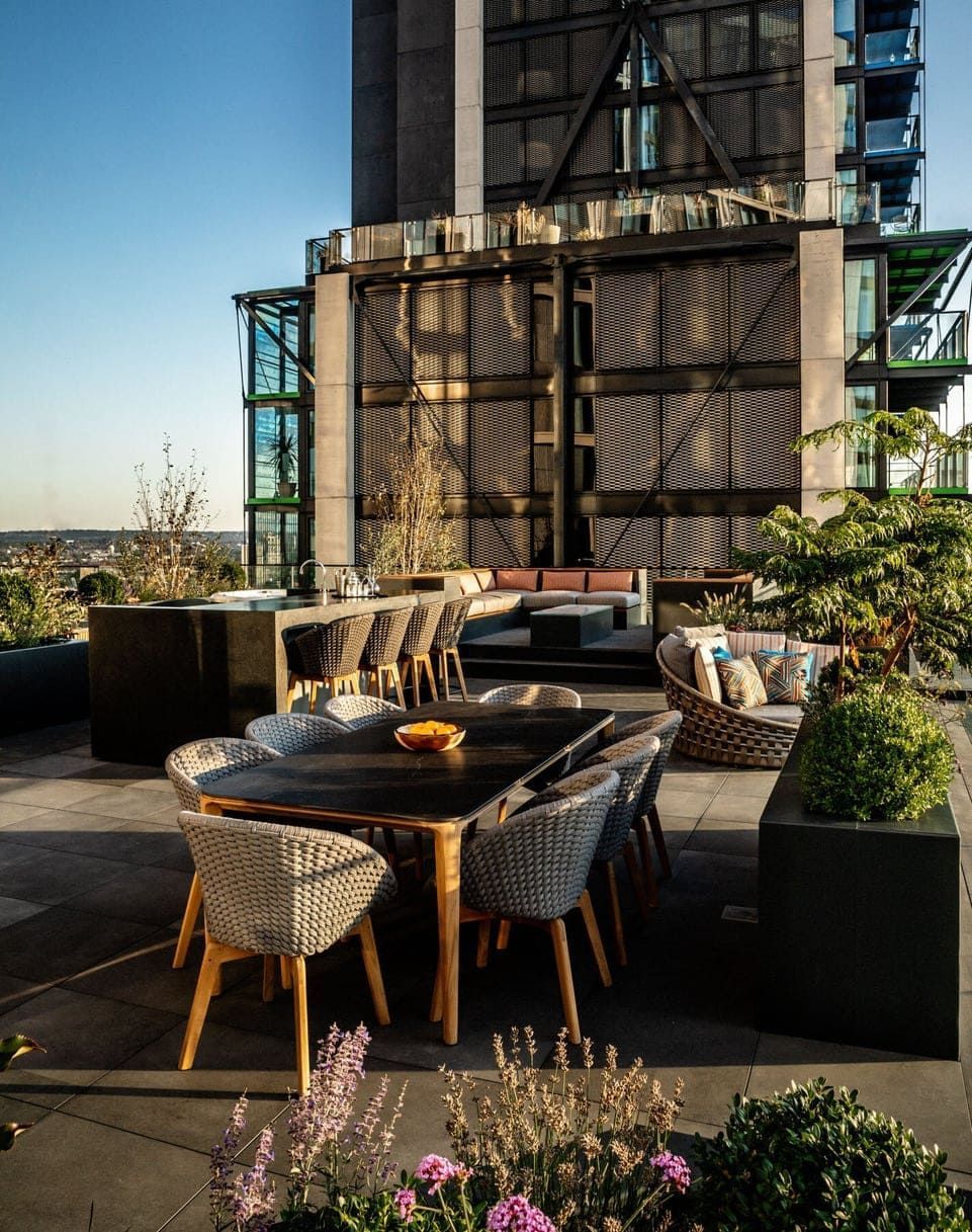 Rooftop patio with dining table and chairs, plush seating, and lush plants against a tall building backdrop.