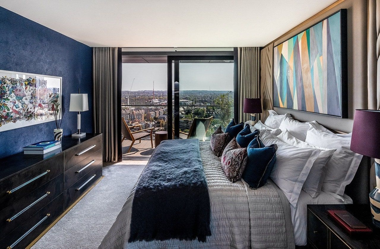 Spacious bedroom with city view; blue textured wall, dark furniture, large bed with colorful pillows.
