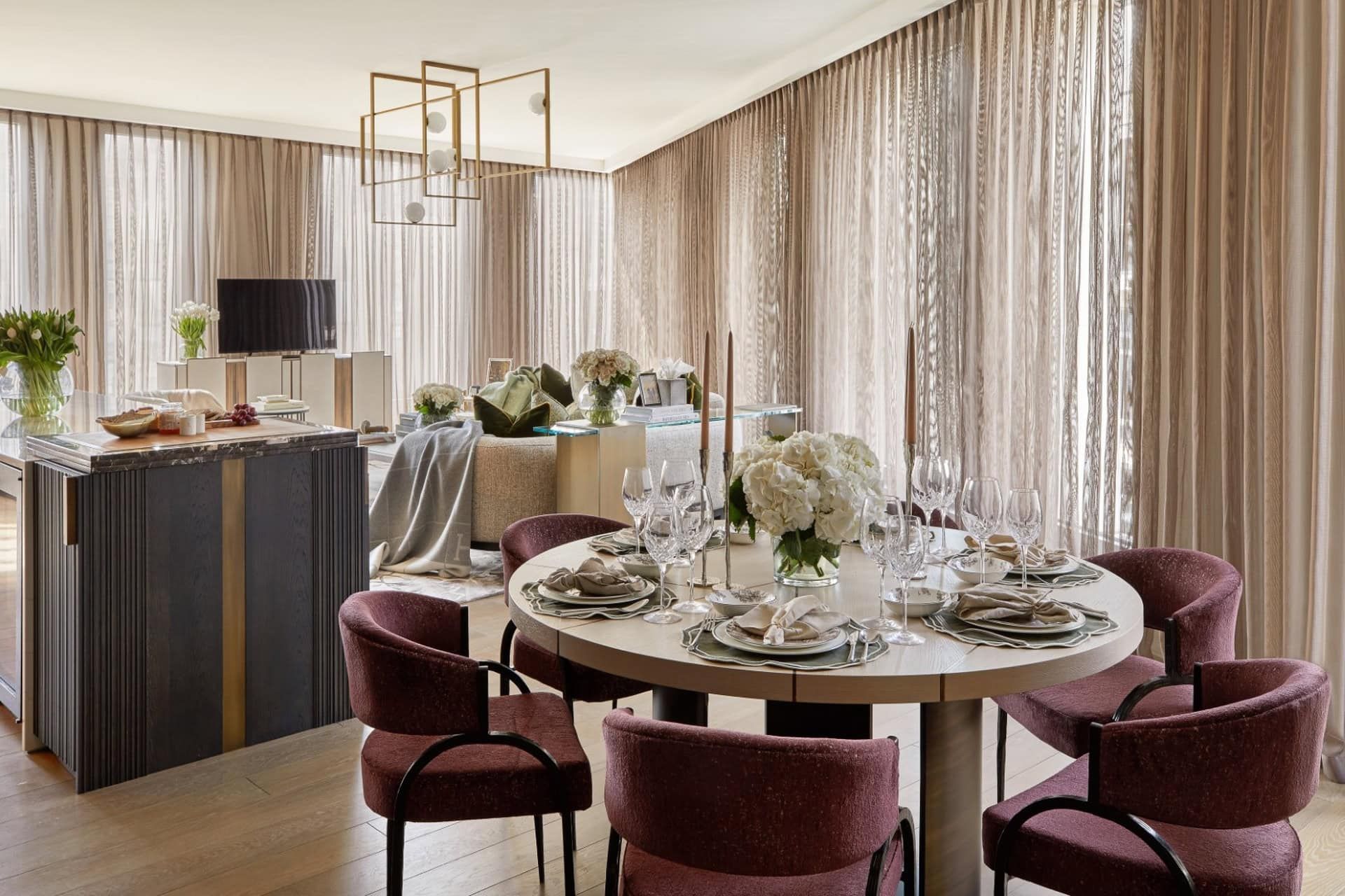 Elegant dining room with round table set for guests, dark red chairs, and tan curtains.