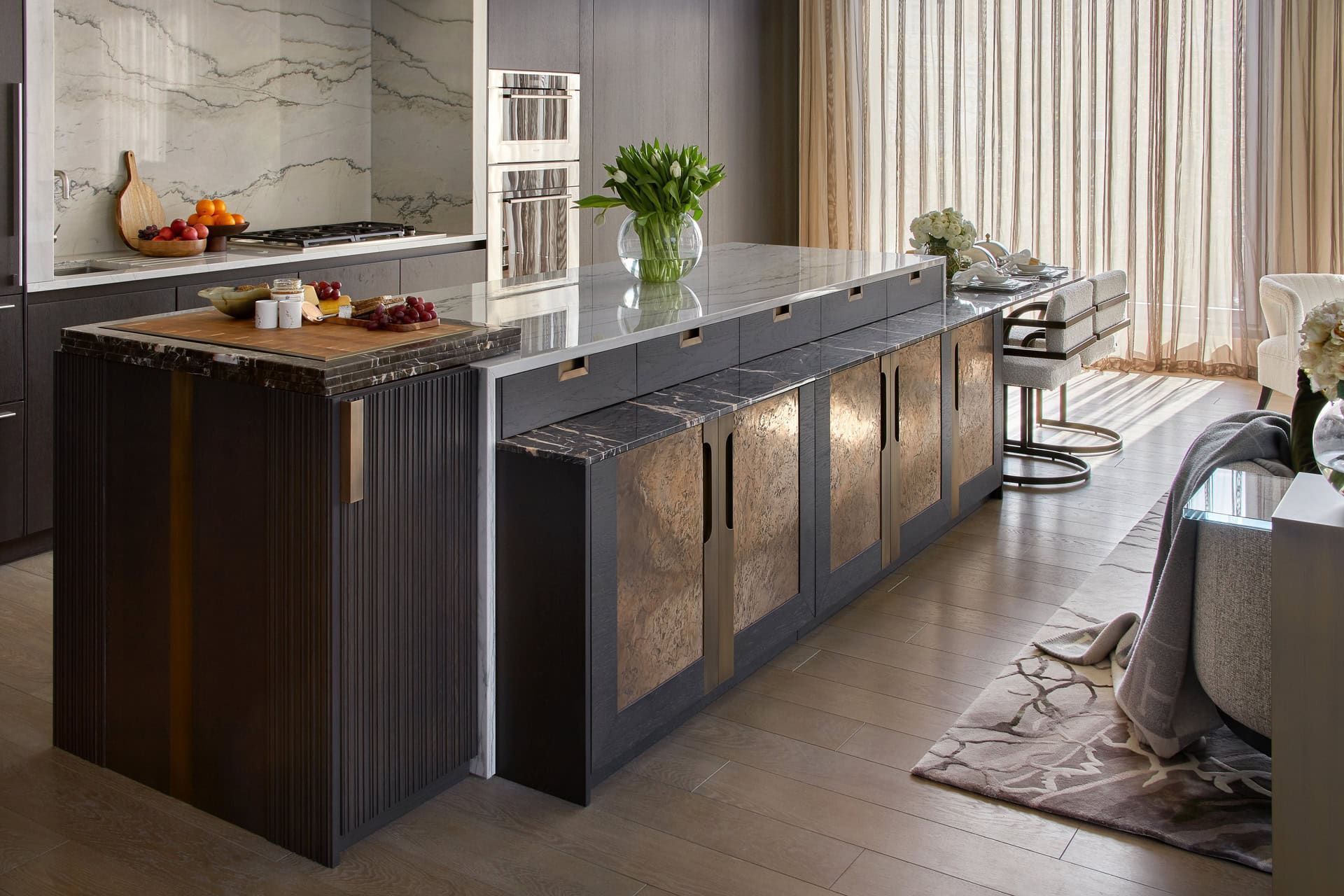 Modern kitchen with island, dark wood, bronze accents, marble countertops, and stools.