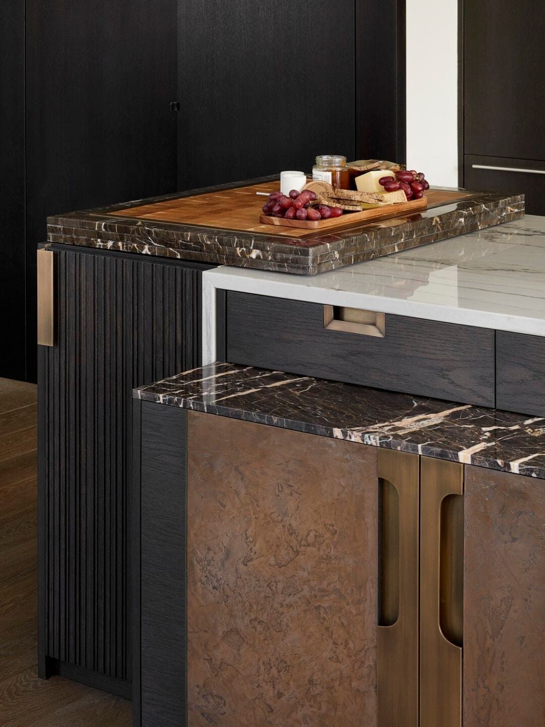 Close-up of a modern kitchen island with wood and stone countertops, food displayed, and bronze cabinet hardware.