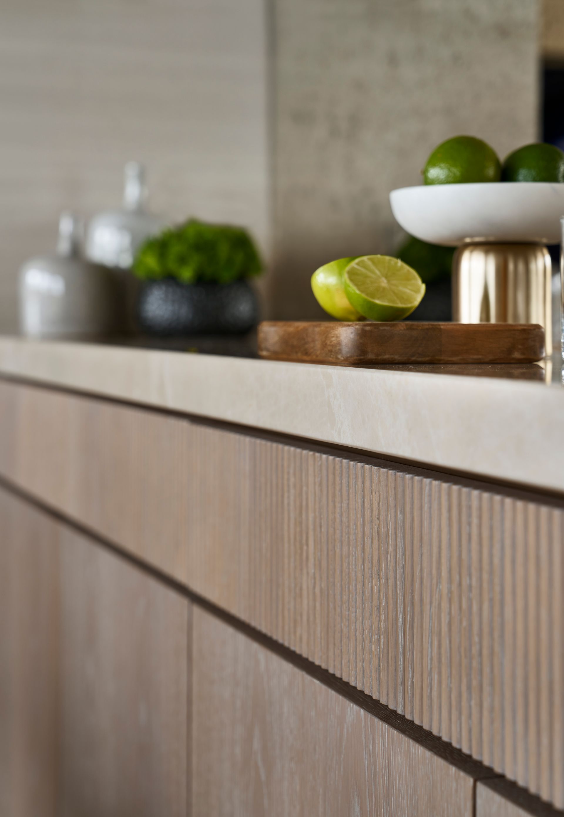 Close-up of a kitchen counter with a wooden cabinet. Lime slices rest on a wooden cutting board, and a bowl of limes sits nearby.