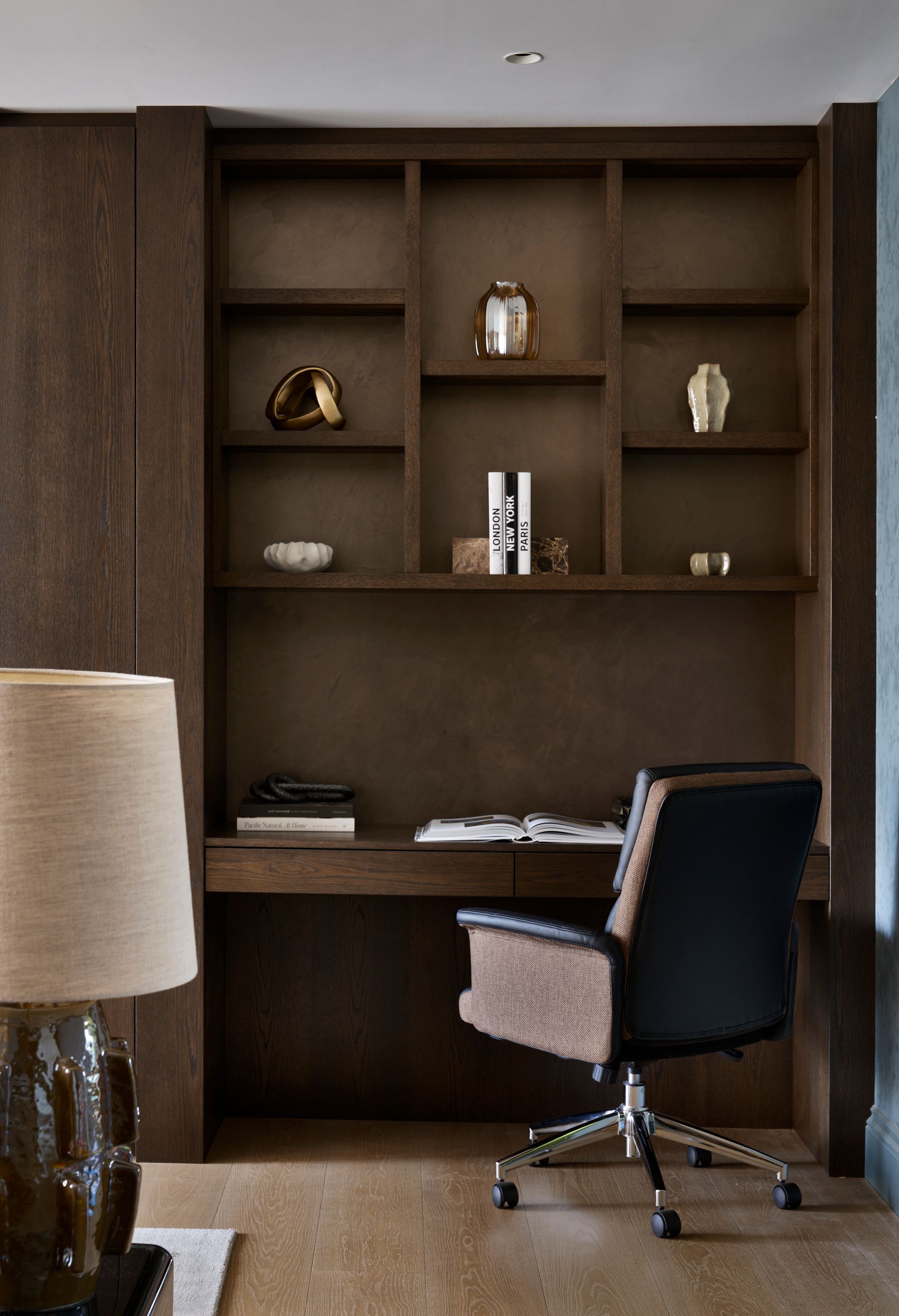 Dark wood home office with built-in desk, shelving, and black office chair; decorated with sculptures and books.
