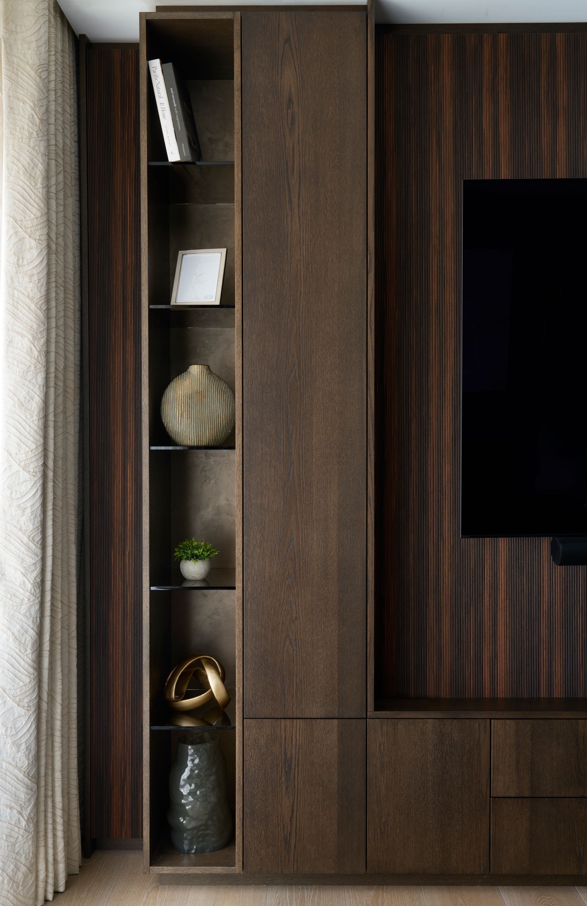 Tall, dark wood cabinet with shelves, holding decorative objects, next to a TV on a dark, wood-paneled wall.