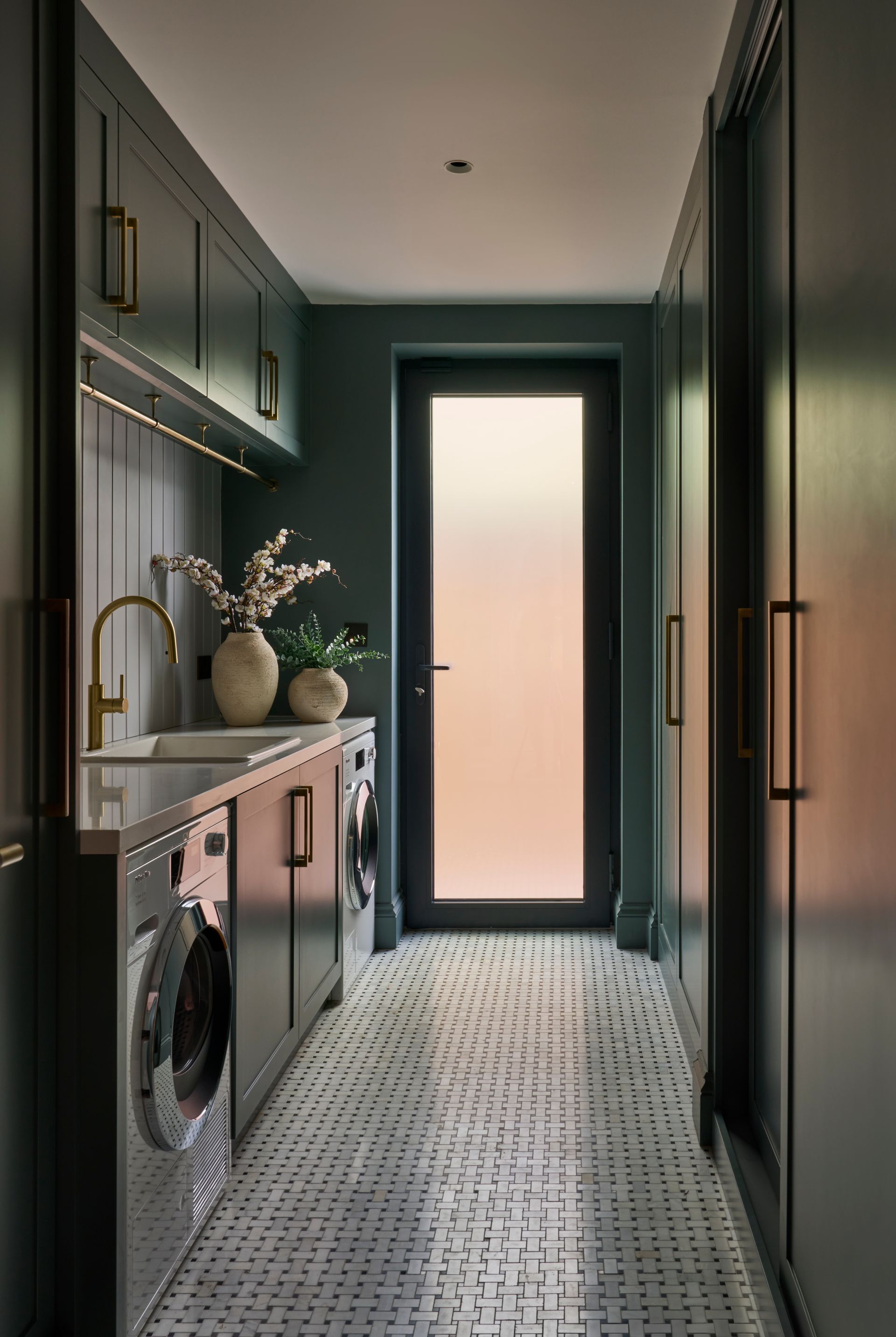 Narrow laundry room with dark green cabinets, white and gray patterned floor, washing machines, and a frosted glass door.