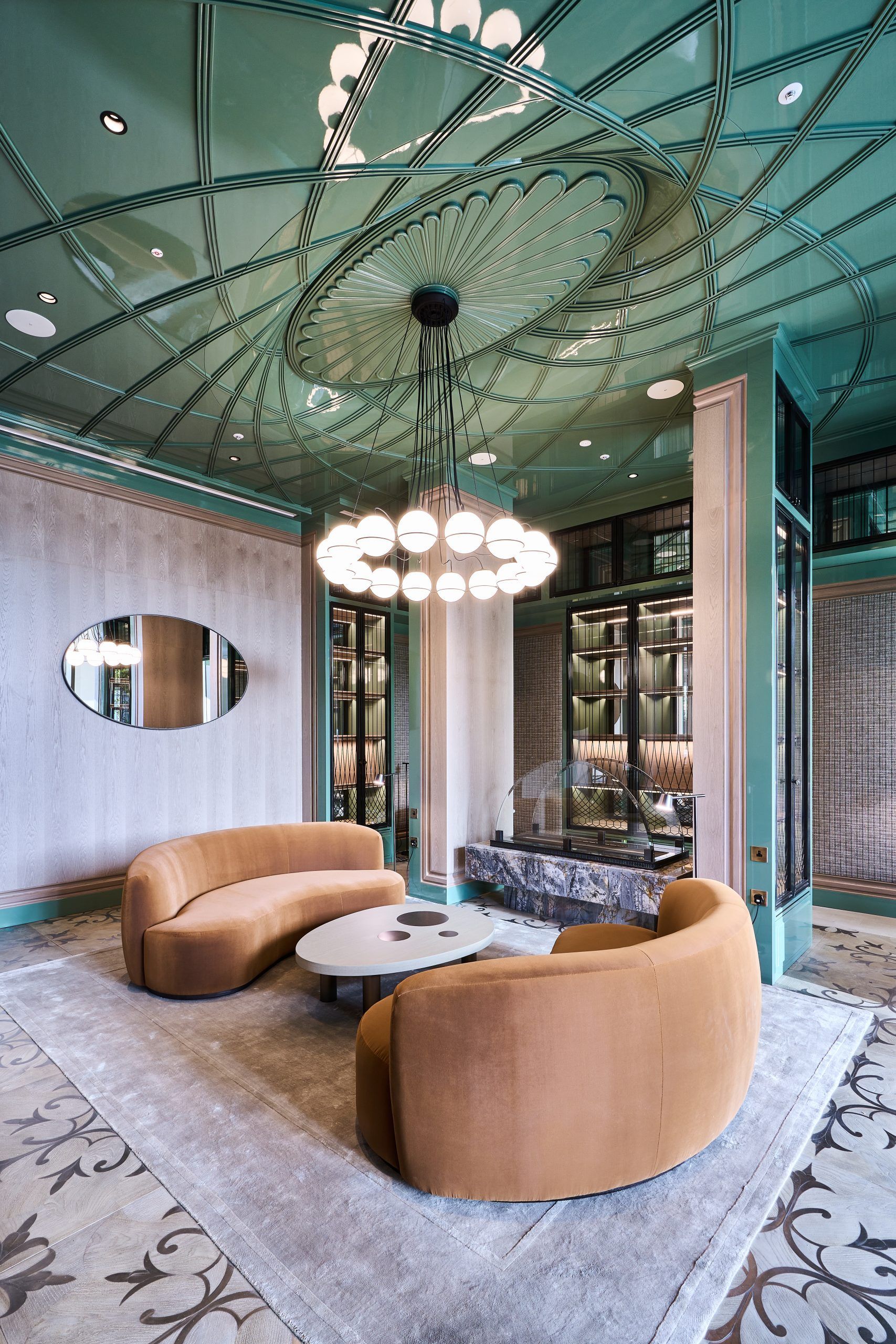 Lounge area with curved orange sofas, round table, and a patterned rug under a mint green ceiling with a chandelier.
