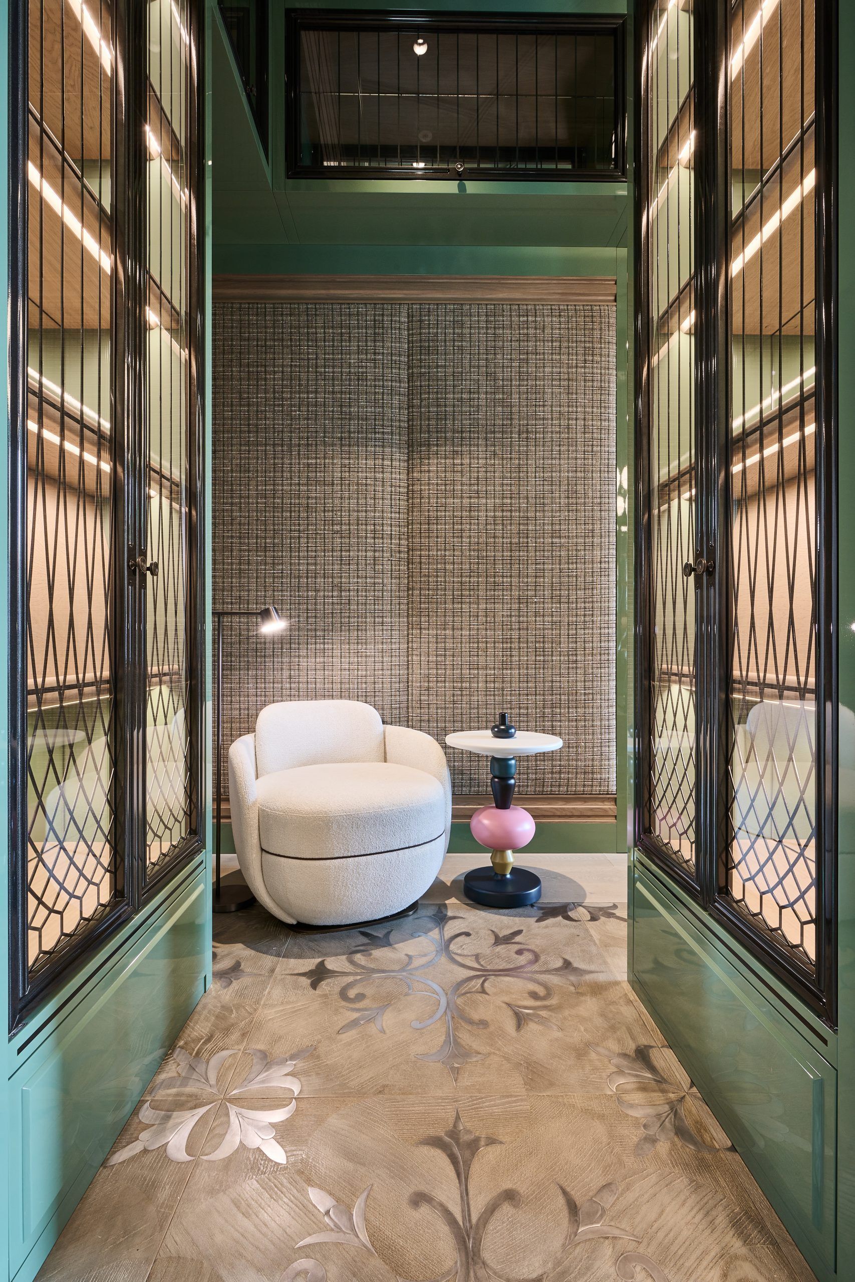 Green hallway with ornate glass doors leading to a room with a cozy white chair and a patterned rug.