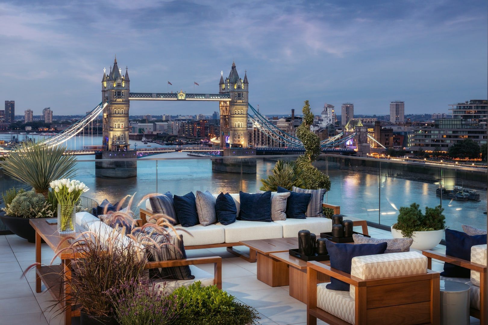 Rooftop lounge with furniture, plants, and view of Tower Bridge in London at dusk.