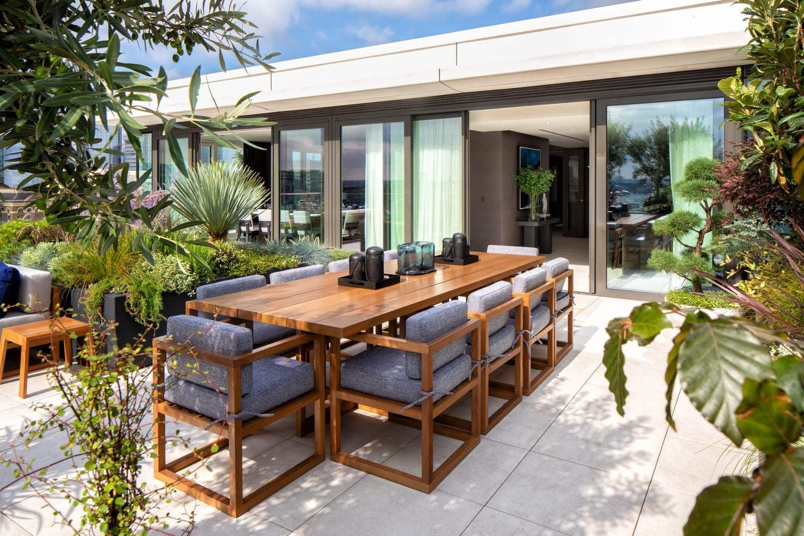 Outdoor dining area with wooden table, blue chairs, and lush greenery, with a building in the background.